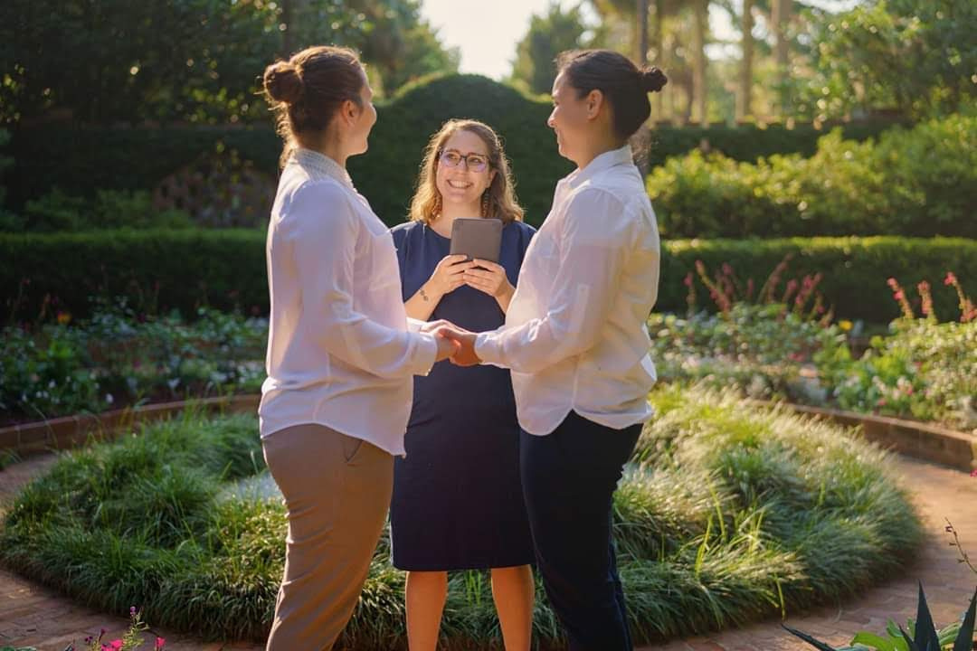 Two women, both with light skin and brown hair in buns and wearing white button-up shirts and khaki slacks facing each other and holding hands. Lily, the Officiant, stands behind in a navy blue dress, holding a book.