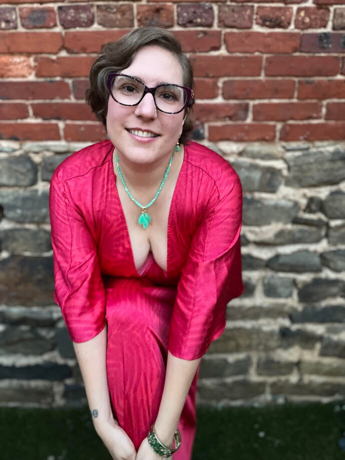 Lily, a petite white nonbinary woman with short dark blonde hair and purple glasses, wearing a hot pink, light aqua beaded necklace and earrings,
Standing in front of an brick wall slightly bent forward, hands clasped in front of one raised knee