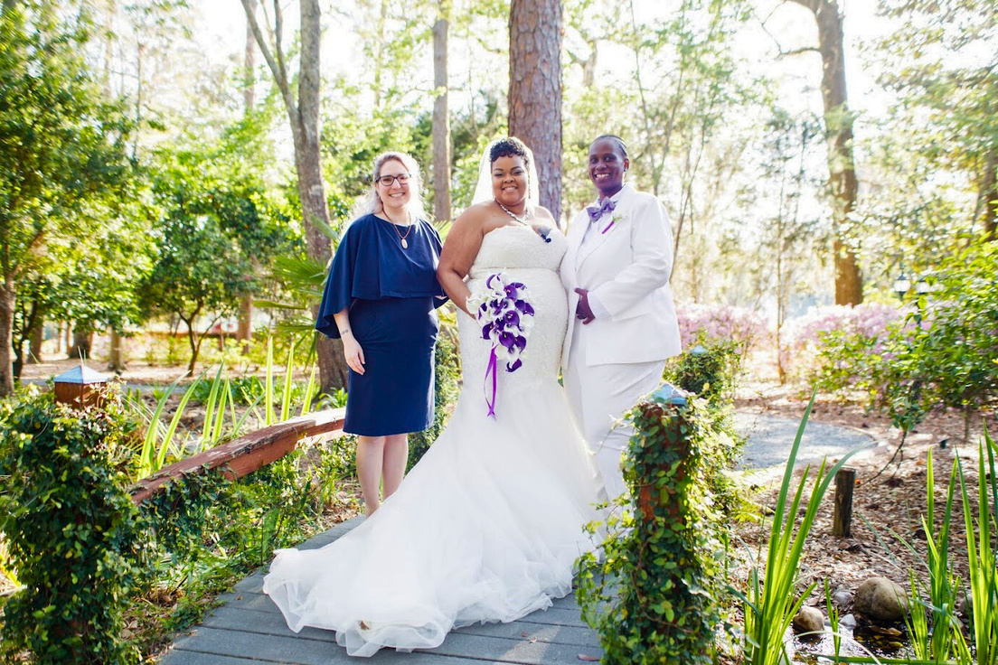 A Black woman with medium skin wearing a chapel length wedding dress and her wife, a Black woman with dark skin wearing a white tuxedo and purple bow tie,  standing on a small wooden bridge. Lily stands to the left of them in a navy blue dress.